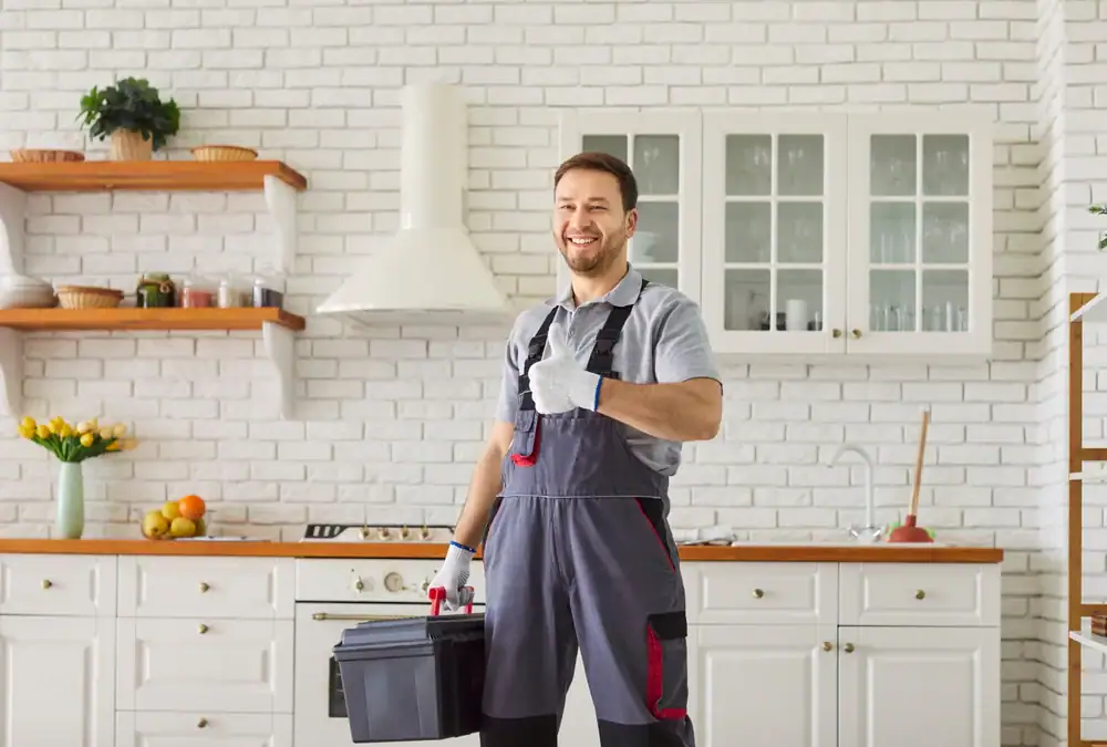 A smiling plumber Pasco County expert in overalls and gloves stands in a bright FL kitchen, holding a toolbox and giving a thumbs-up. The space features white cabinets, a brick backsplash, and various utensils on the countertop.