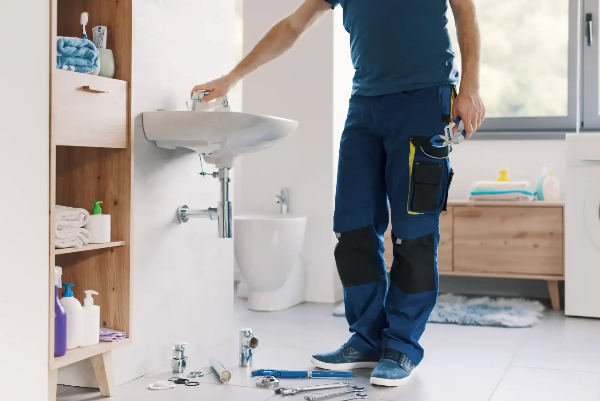 A plumber Pasco County in work clothes repairs a bathroom sink, holding a pipe fitting. Tools and plumbing parts are spread on the floor, with bathroom shelves and a washing machine visible in the background in FL.