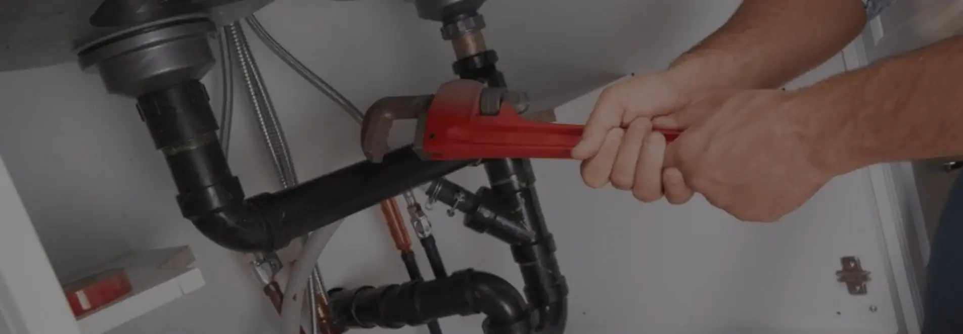 A plumber Pasco County, FL uses a red pipe wrench to tighten black plumbing pipes under a sink.