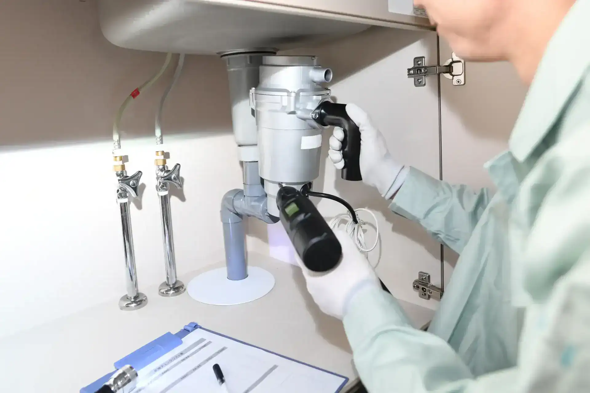 A plumber in Pasco County, FL, wearing gloves inspects a garbage disposal unit under a kitchen sink with a flashlight. Nearby are pipes, valves, and a clipboard with a pen on the countertop.