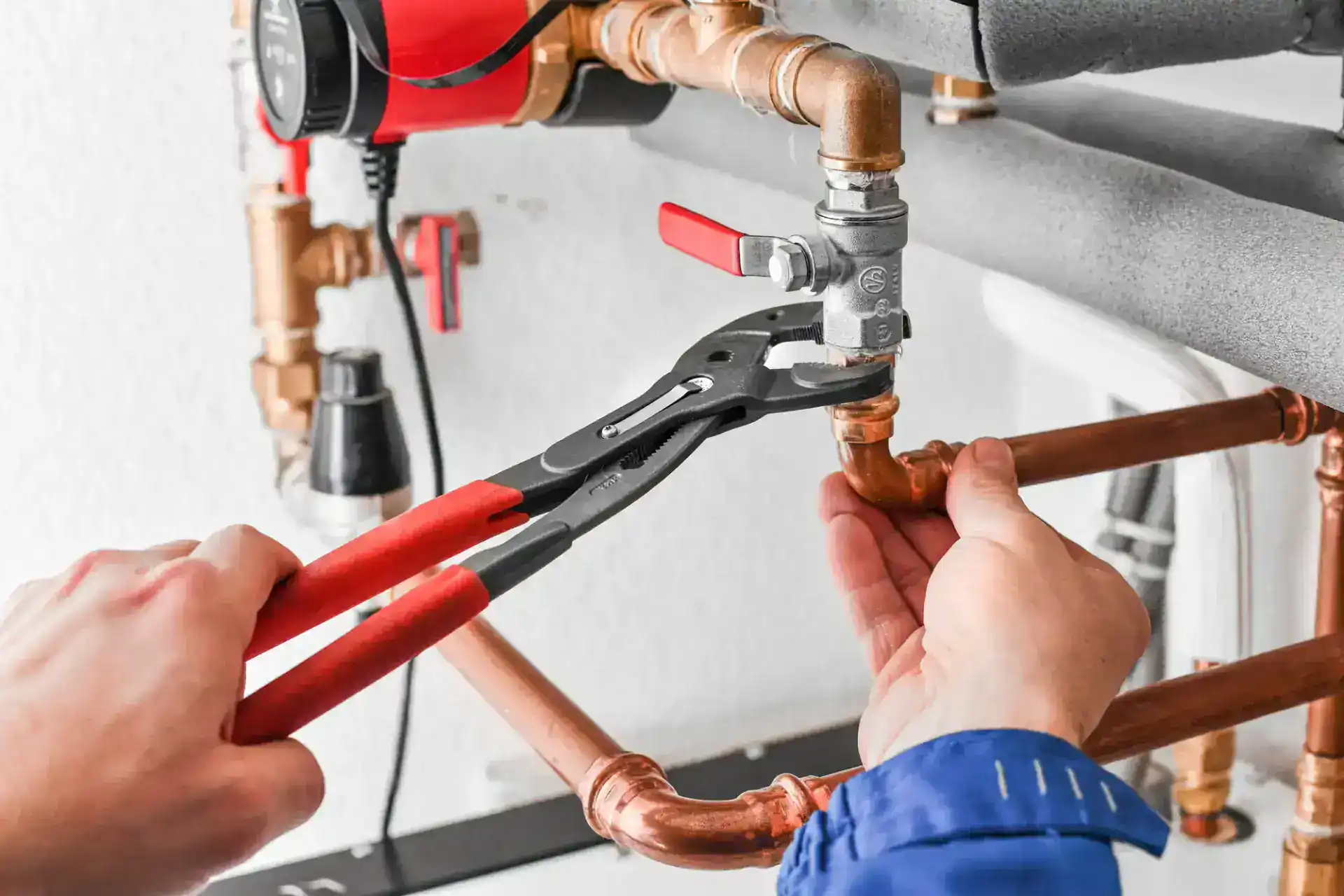 Hands using pliers to tighten a valve on copper plumbing pipes beside a residential water heater system.