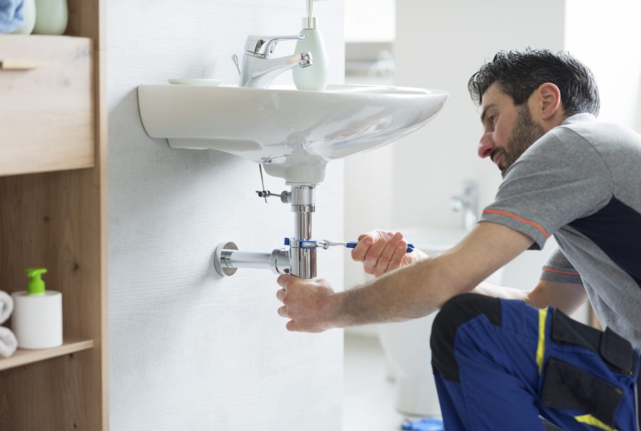 A man kneels fixing a bathroom sink pipe with a wrench under the basin in a bright bathroom.