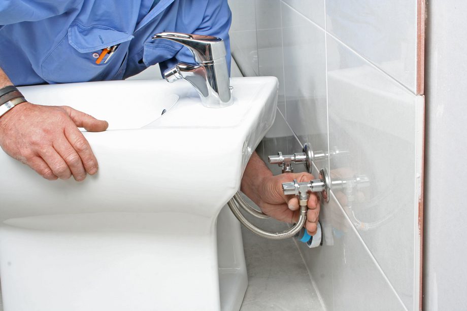 A person repairs metal pipes under a white sink with a silver faucet, shown against tiled bathroom walls.