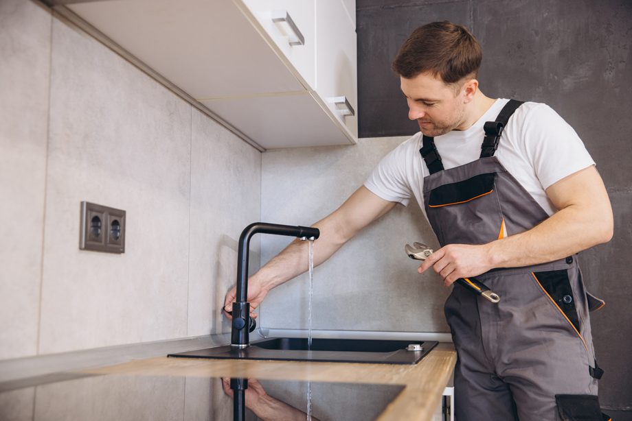 A plumber in gray overalls repairs a kitchen faucet with water running, holding a wrench in one hand.