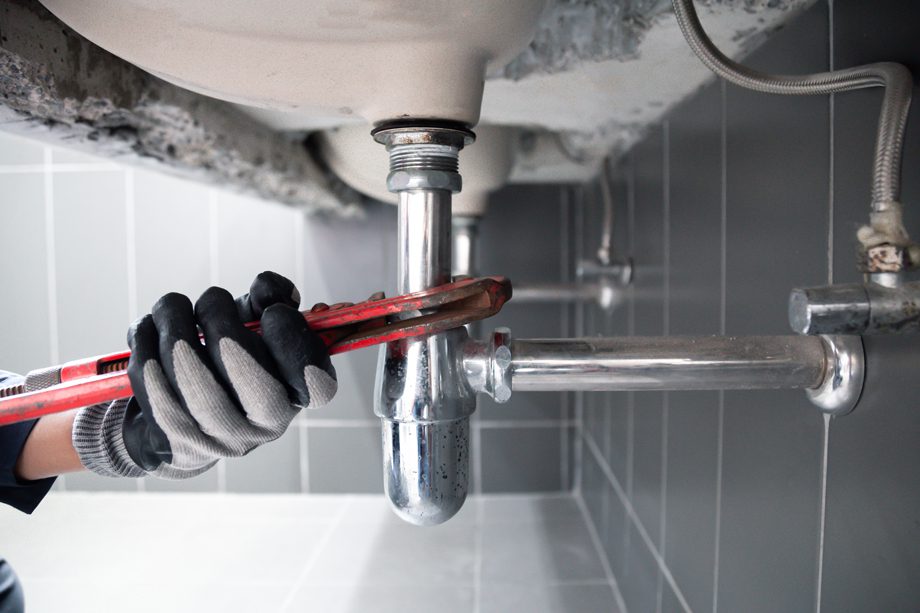 A person wearing gloves tightens a metal pipe under a bathroom sink using a wrench.
