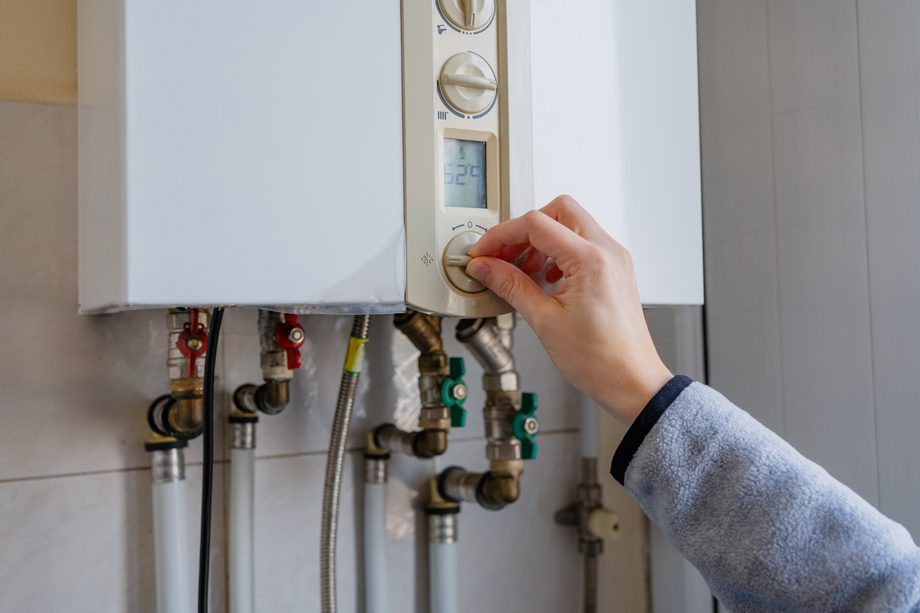 A person adjusts a gas water heater&rsquo;s temperature dial, with pipes visible beneath the wall-mounted unit.