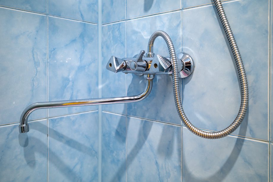 A chrome shower faucet and hose are mounted on a blue tiled bathroom wall in the corner.