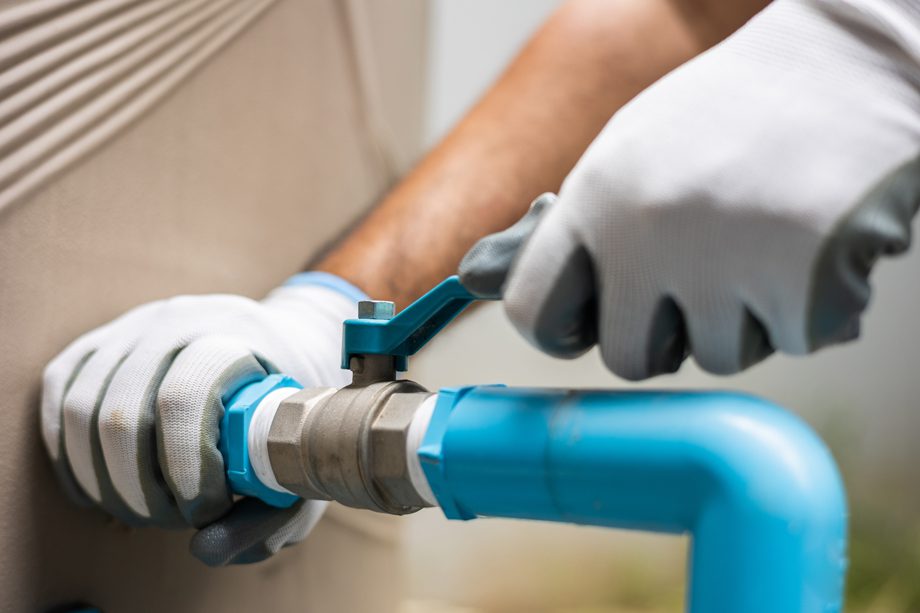 A plumber&rsquo;s gloved hands use a wrench to tighten a blue water pipe for leak repair or installation.