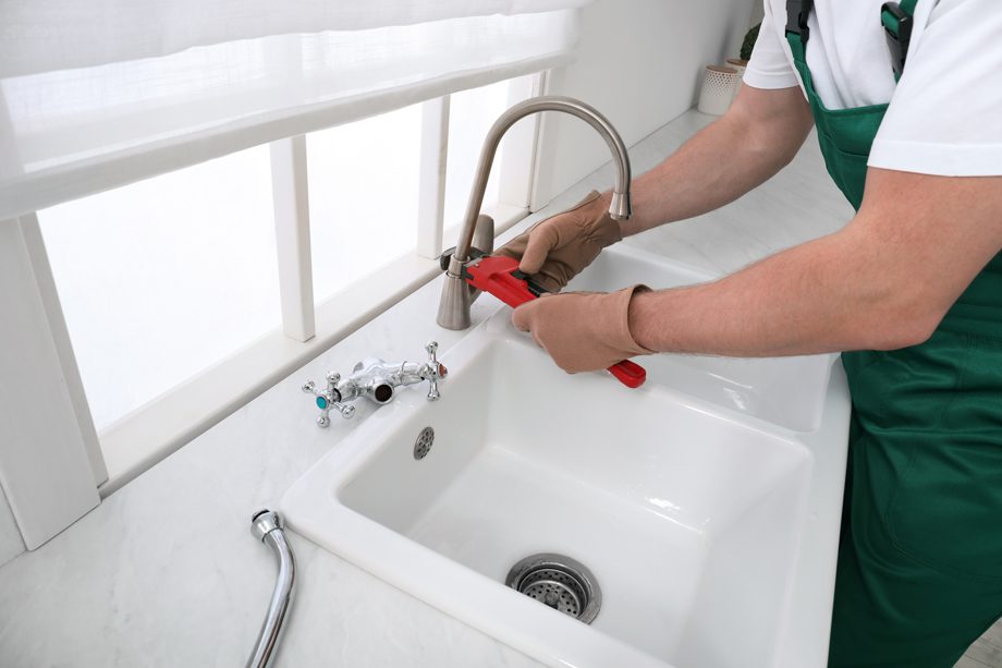 A person in green overalls uses a red pipe wrench to fix a kitchen sink faucet.