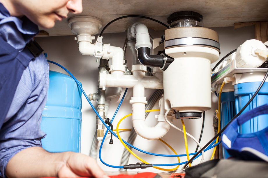 Person inspecting pipes and water filtration system under a sink with visible tubes and plumbing connections.