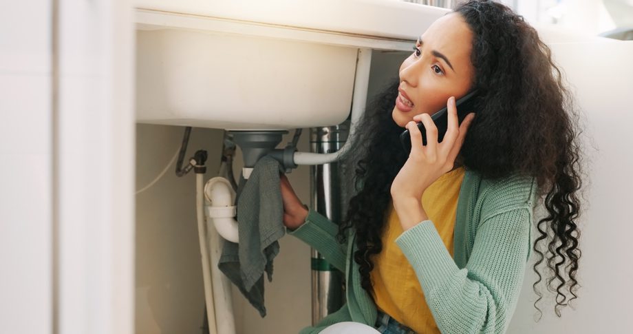 Concerned woman under sink holds cloth and speaks on phone, possibly calling for plumbing help or advice.