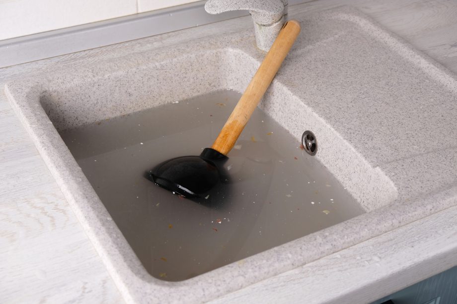 A plunger sits in a clogged kitchen sink surrounded by dirty water and food debris.